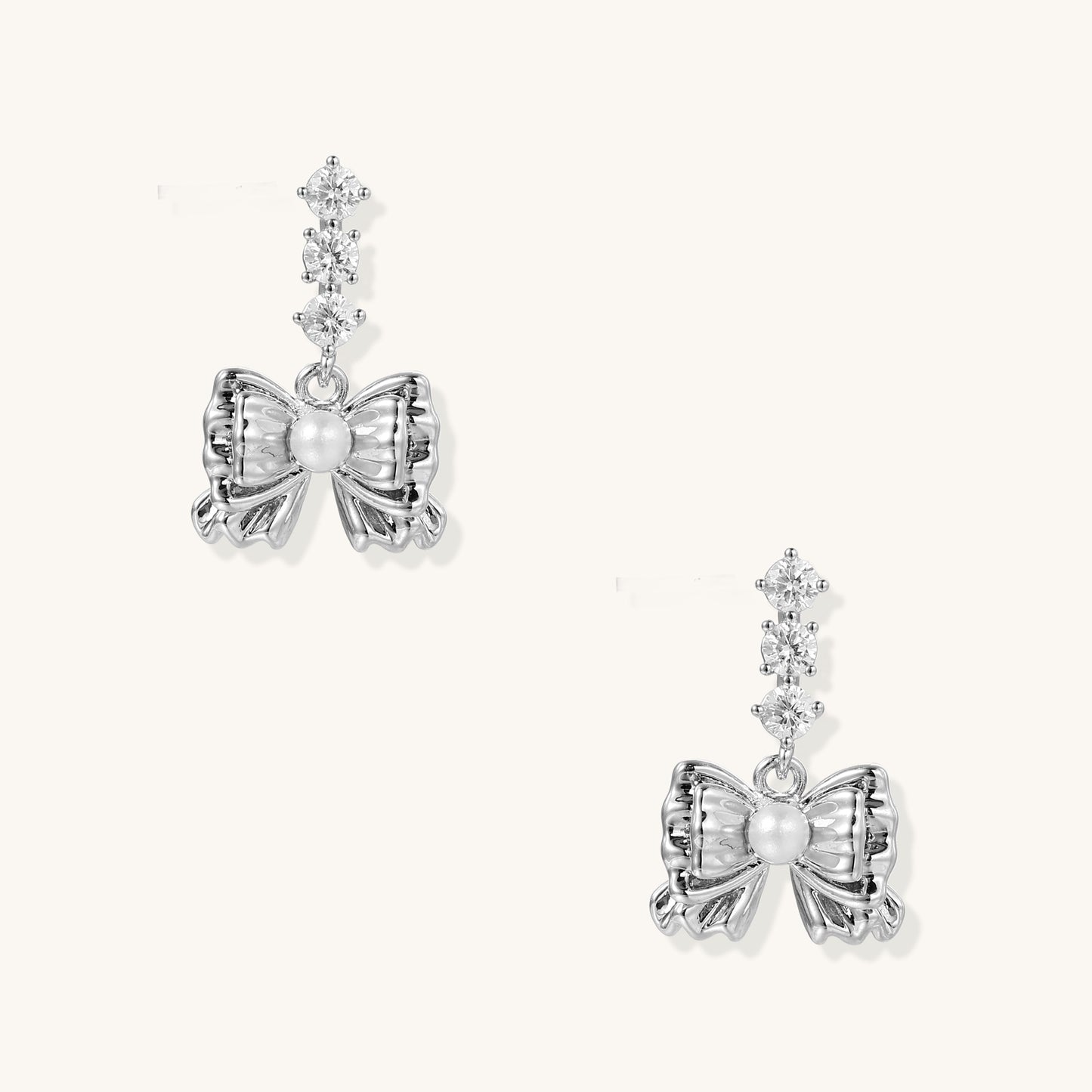 Delicate gold-plated bow drop earrings with three zirconia crystals and center pearl on white background.