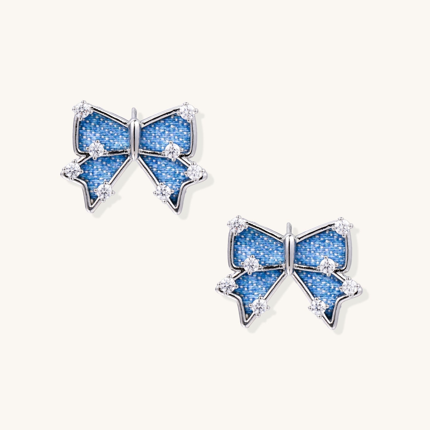 Pair of bow-shaped stud earrings featuring blue denim fabric with white stitching and gold-plated crystal accents