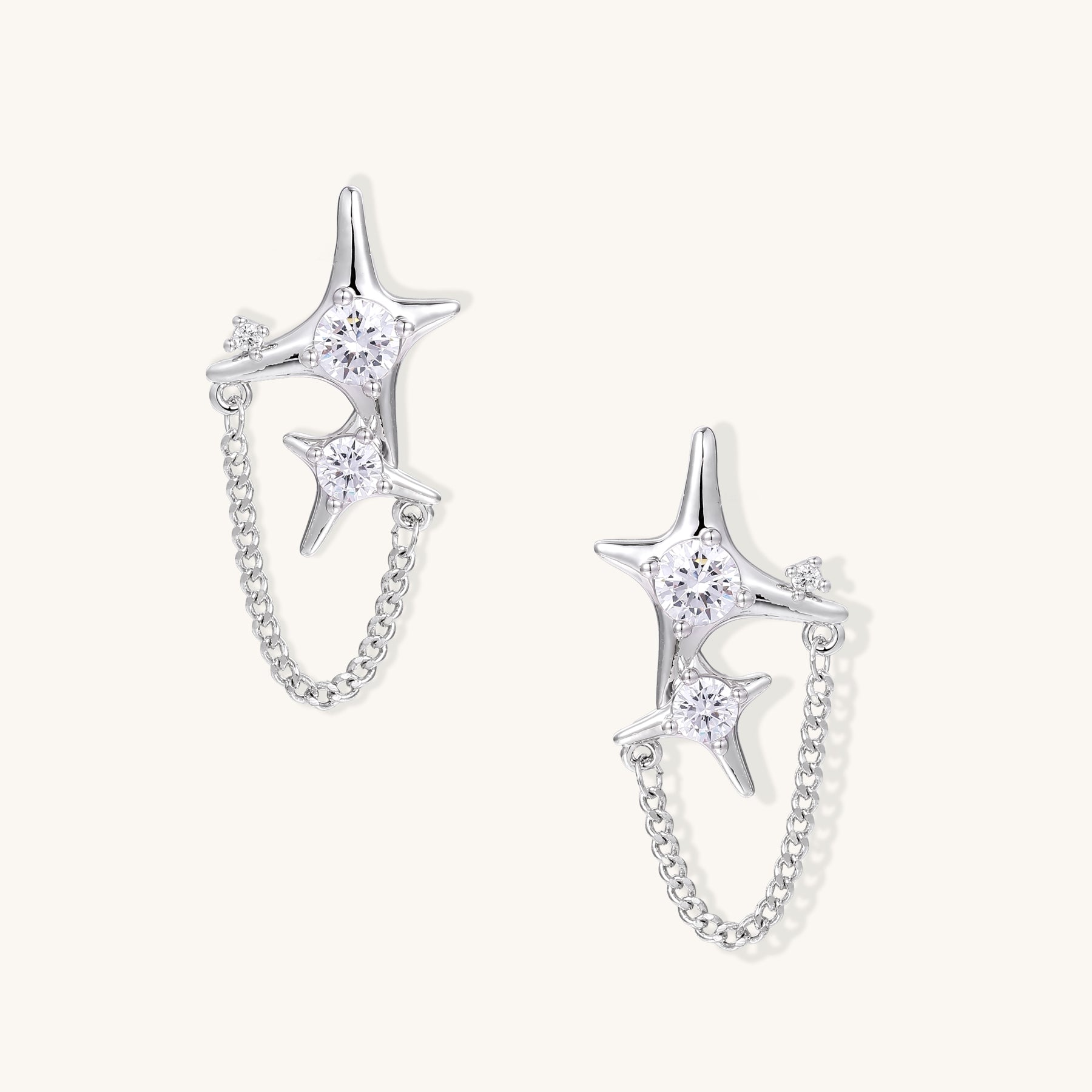 Delicate gold-plated star earrings with sparkling zirconia crystals connected by fine chains on cream background.